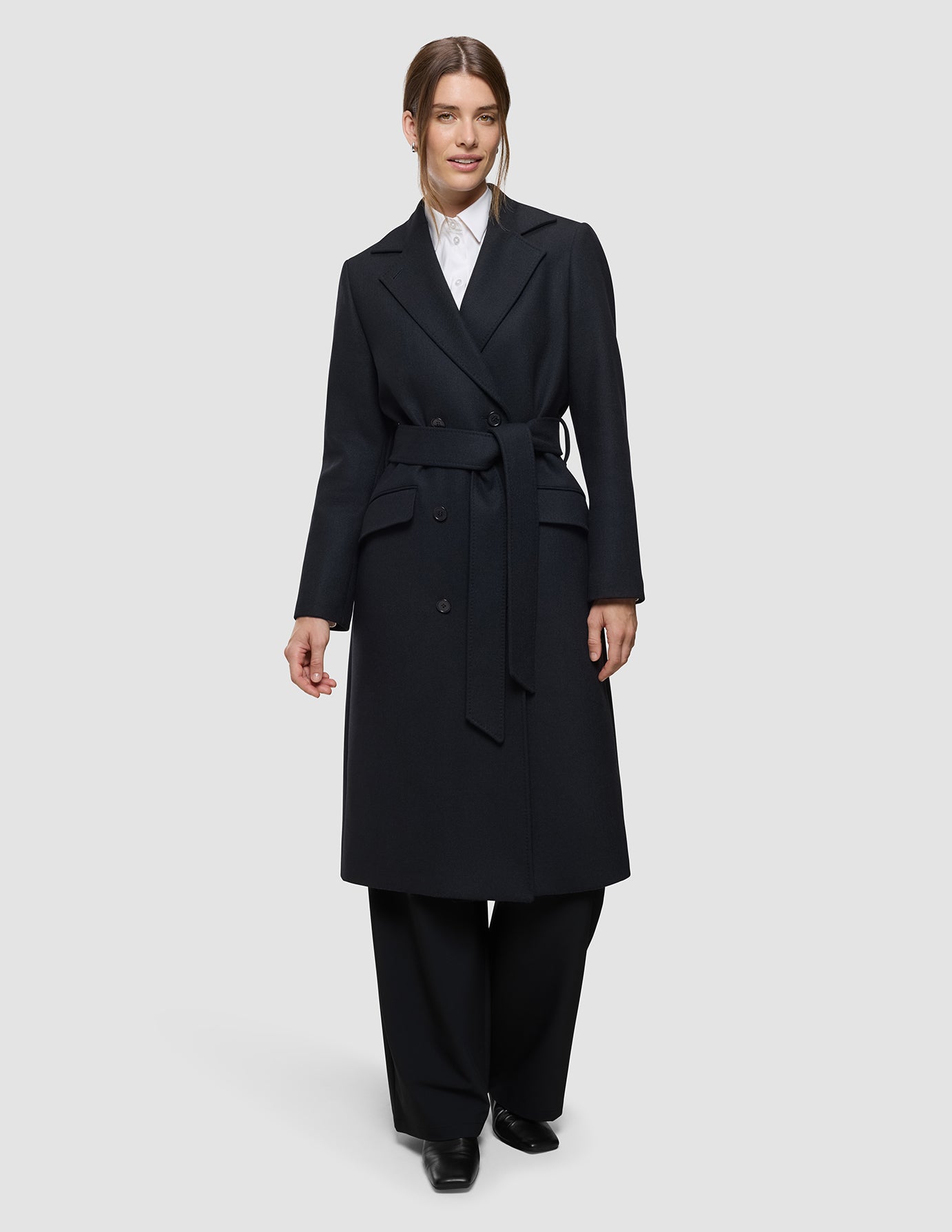 En person iført en sort uldfrakke til kvinder med bælte over en hvid skjorte med krave og sorte bukser i et klassisk design.